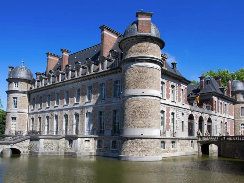 Beloeil Castle in Belgium, showcasing its elegant façade and grand architecture. The castle features a classic moat and is set against a backdrop of lush, green trees and a clear sky.