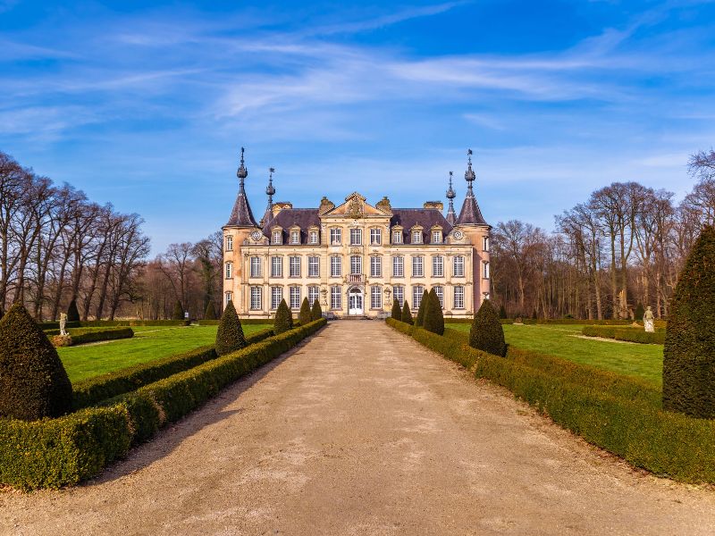 Castle of Poeke displaying its classic medieval architecture with a grand stone façade, tall towers, and elegant turrets. The castle is surrounded by expansive gardens and a reflective moat, set against a peaceful countryside backdrop.