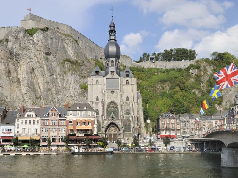 Citadel of Dinant perched on a rocky hill overlooking the town, with a dramatic cliffside backdrop and the Meuse River flowing below. The historic fortress features stone walls and a distinctive tower against a scenic sky.