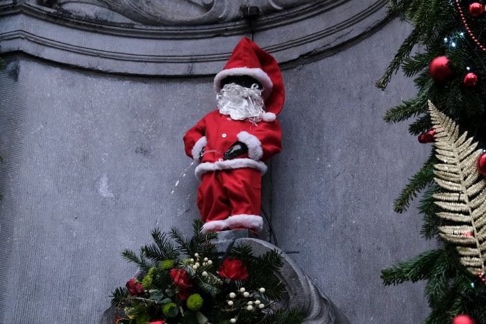 The Manneken Pis statue in Brussels is dressed in a festive Santa Claus outfit, complete with a red suit, white fur trim, and a curly white beard. Water continues to flow from the statue as part of its famous fountain display. Surrounding the statue are Christmas decorations, including garlands, red ornaments, and a floral arrangement, enhancing the holiday theme.