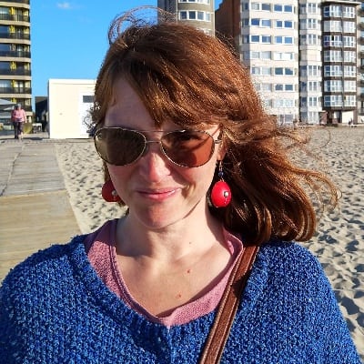 headshot of a white woman with medium long brown hair, a blue sweater and pilot-style sunglasses at the beach