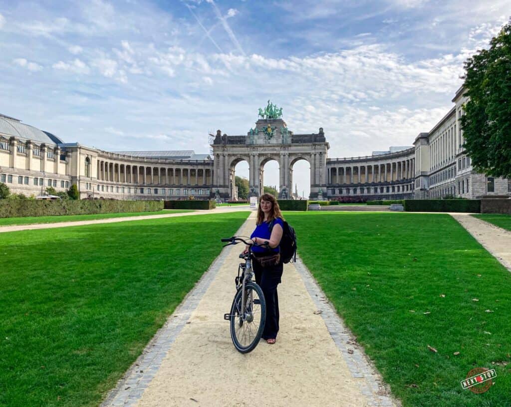 A woman with a bike stands in front of the grand arches of Parc du Cinquantenaire on a sunny day."