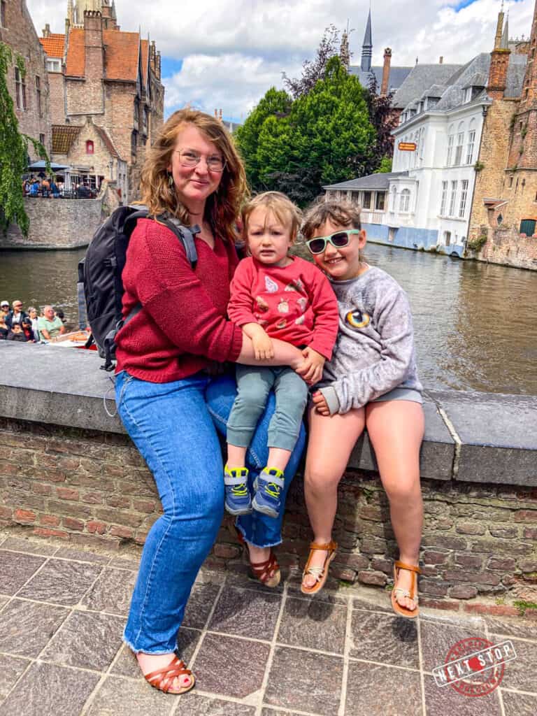 Mom with two kids posing in the scenic canal views from Rosary Quay in Bruges, Belgium, showcasing charming medieval buildings with red-tiled roofs reflected in the calm water.