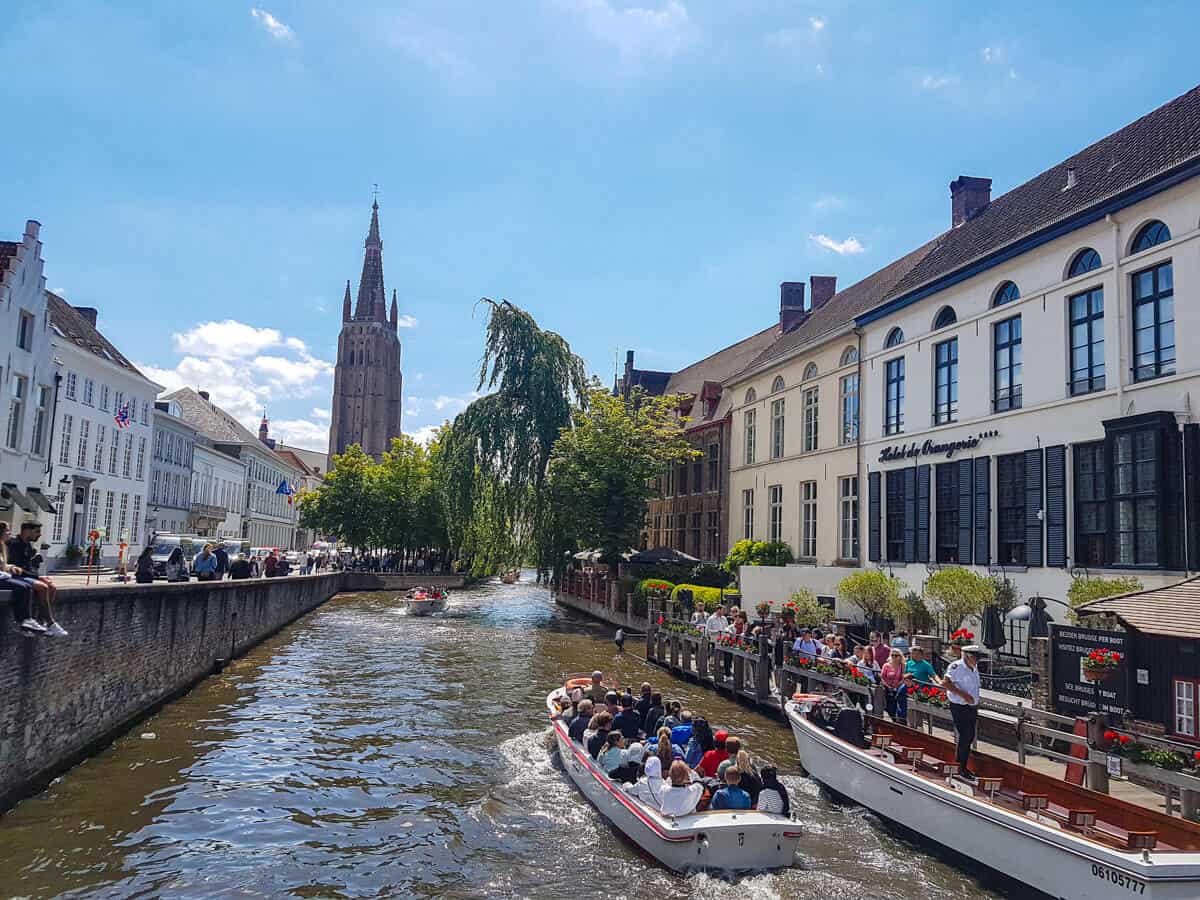 Canal in Bruges with a tour boat and a church tower in the distance - all under a clear blue sky.