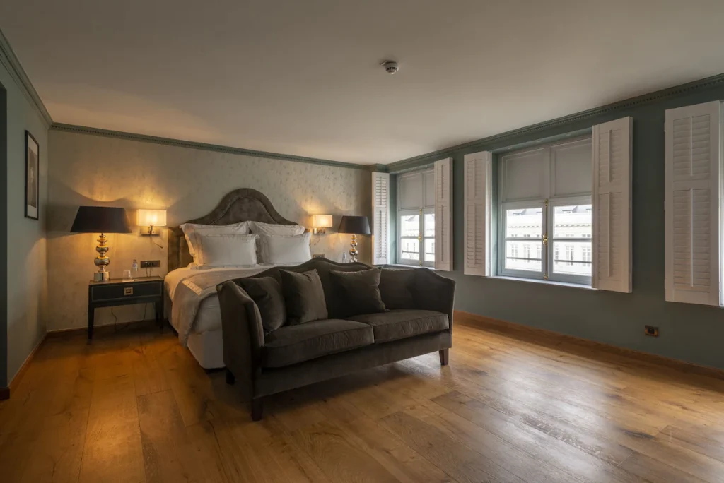 A green bedroom in one of the best hotels in Brussels City Centre with wooden floor, bed, couch, bedside tables with lamps, and windows