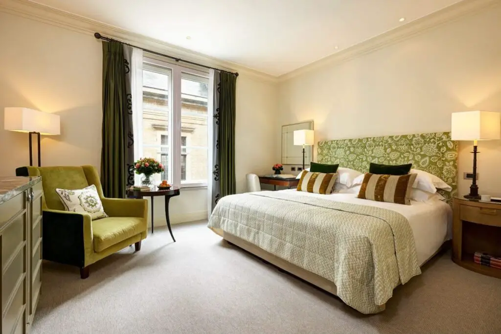 A carpeted bedroom with green bed frame, white bed, tables, lamps, green chair, counter, white windows, and floor-to-ceiling dark green curtains