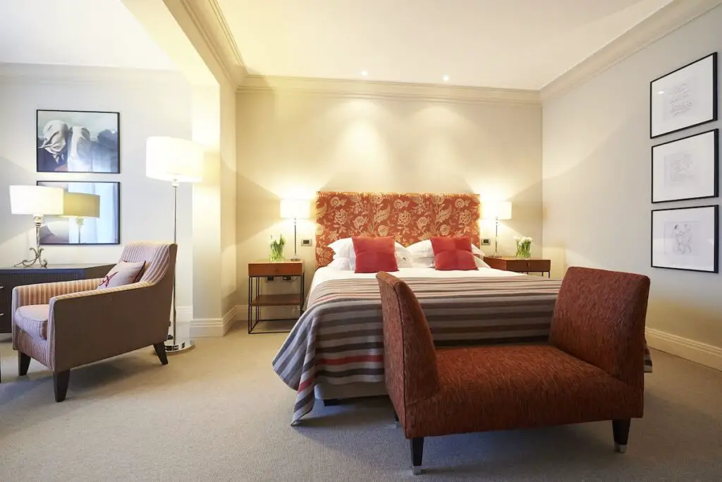 A carpeted room with a red bed frame, red chairs, bedside tables, lamps, and framed art pieces on the wall