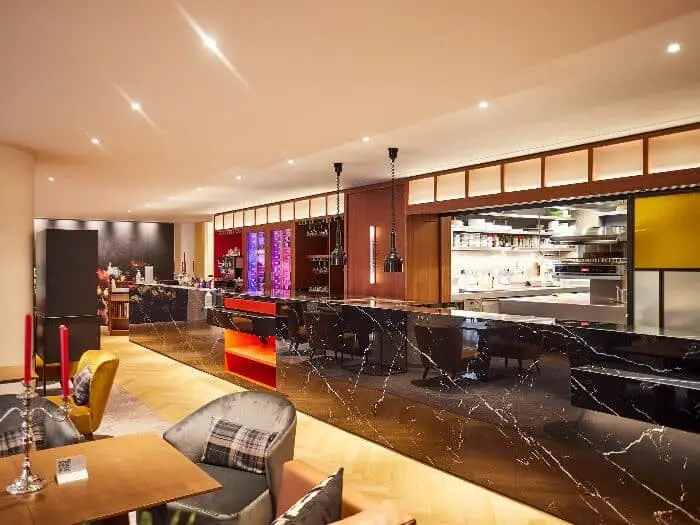 A Brussels City Centre hotel restaurant with a dark marble bar counter, tables, chairs, refrigerators, wine glasses, and a view of the kitchen