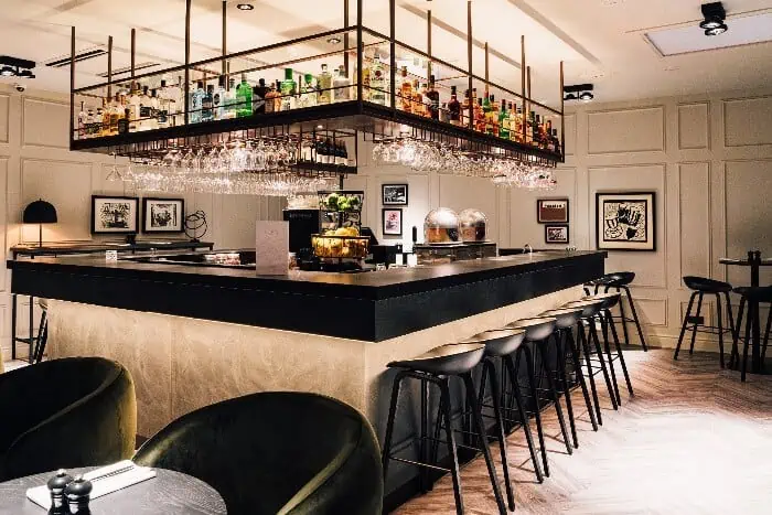 A luxurious restaurant bar with tables with chairs, tall chairs at the counter, and with bottles of drinks and drinking glasses displayed overhead the bar