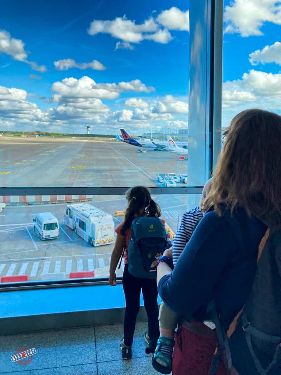 A photo of 3 people near a full-length window with an outside view the sky with clouds above, and parked airplanes and other transportations on the ground.