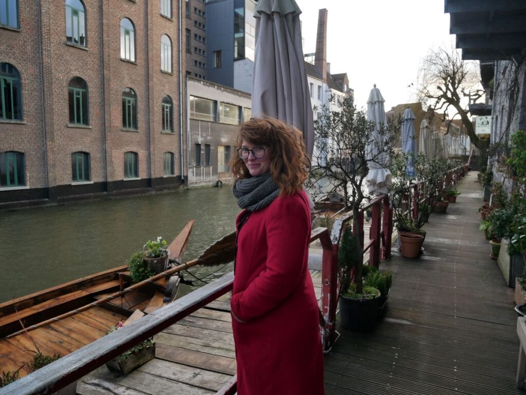 Babs wearing a red coat standing at the side of the river Leie in Ghent.