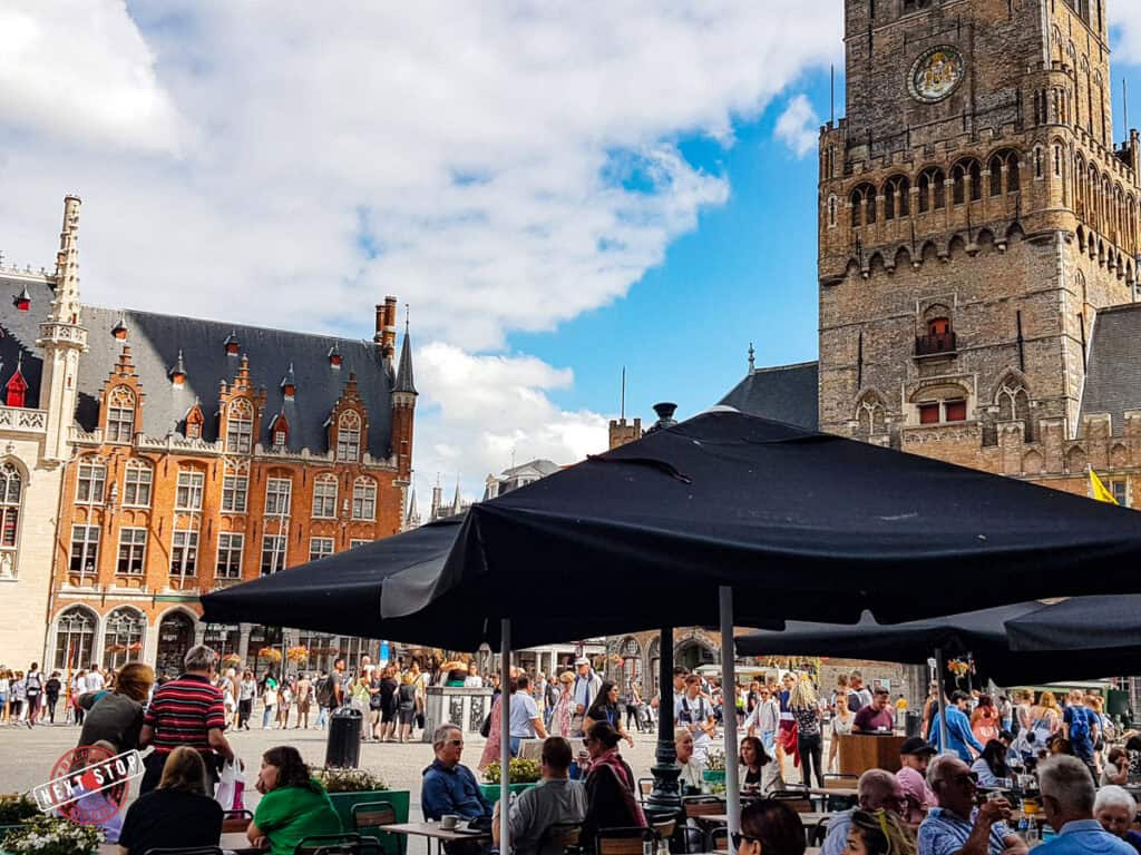 Lots of people having a drink at a terrace on the Market Square and restaurants in Bruges with the Belfry in the back.