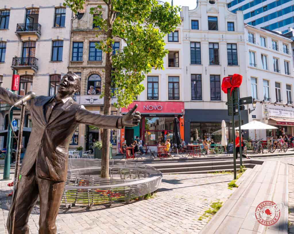 A bronze statue of Jacques Brel, arms wide open as if mid-performance, stands proudly in Place de la Vieille Halle aux Blรฉs, Brussels. Surrounding the statue are charming cafรฉ terraces, cobblestone streets, and historic buildings under a bright blue sky.
