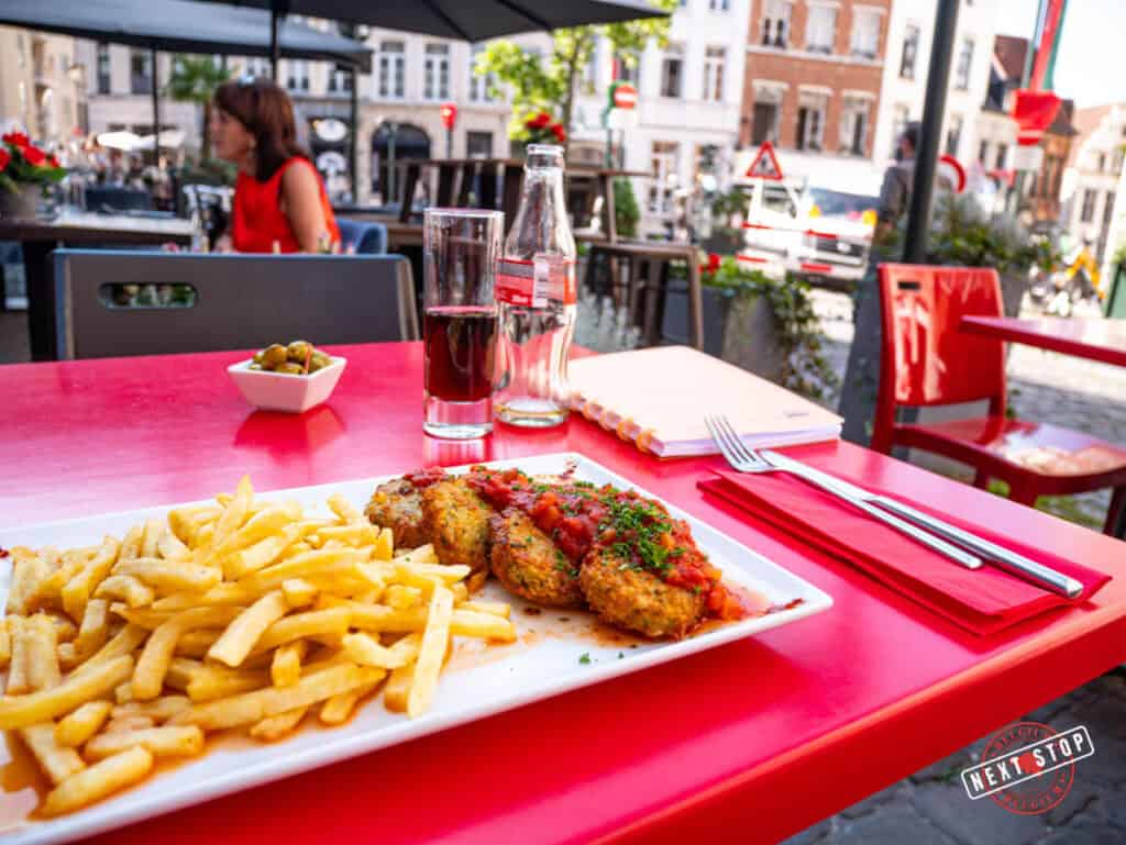 A close-up of a meal served outdoors on a red table at a Brussels restaurant. The dish features crispy golden fries, breaded croquettes with tomato sauce, and a garnish of fresh herbs. A glass of soda and a small bowl of olives are also visible.