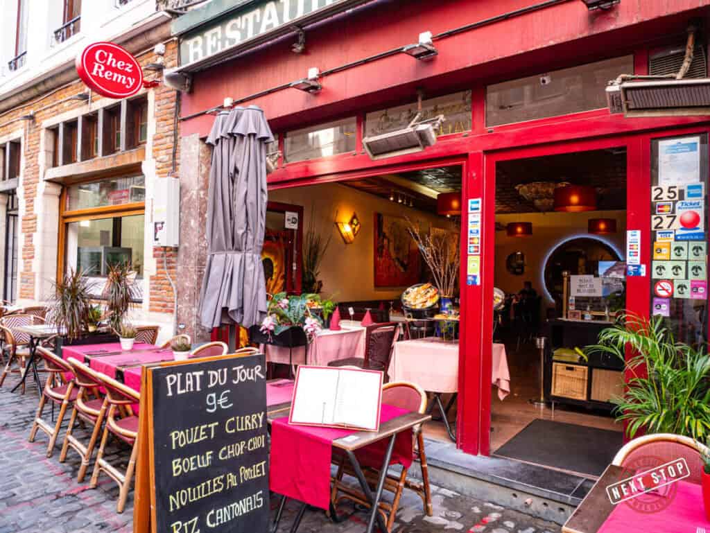 The entrance of Chez Remy restaurant on Rue des Bouchers in Brussels, featuring outdoor seating with red tablecloths and a menu board displaying the daily specials.