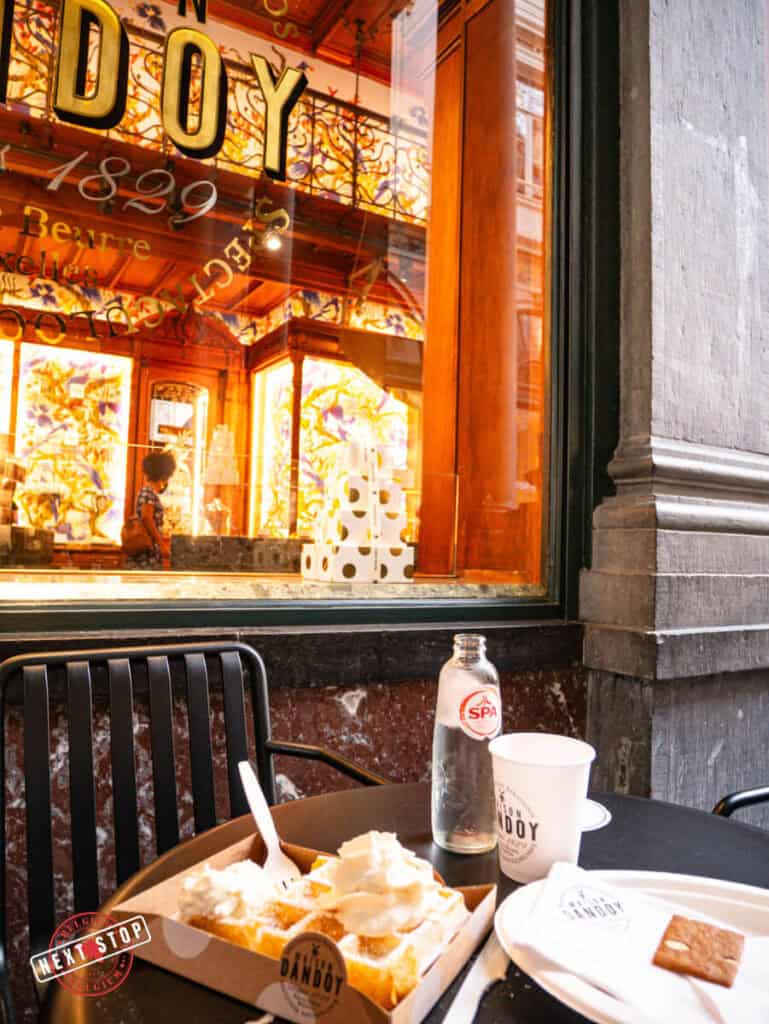 A table outside Maison Dandoy at the St. Hubertus Galleries in Brussels, featuring a box of Belgian waffles topped with whipped cream and a biscuit alongside drinks.