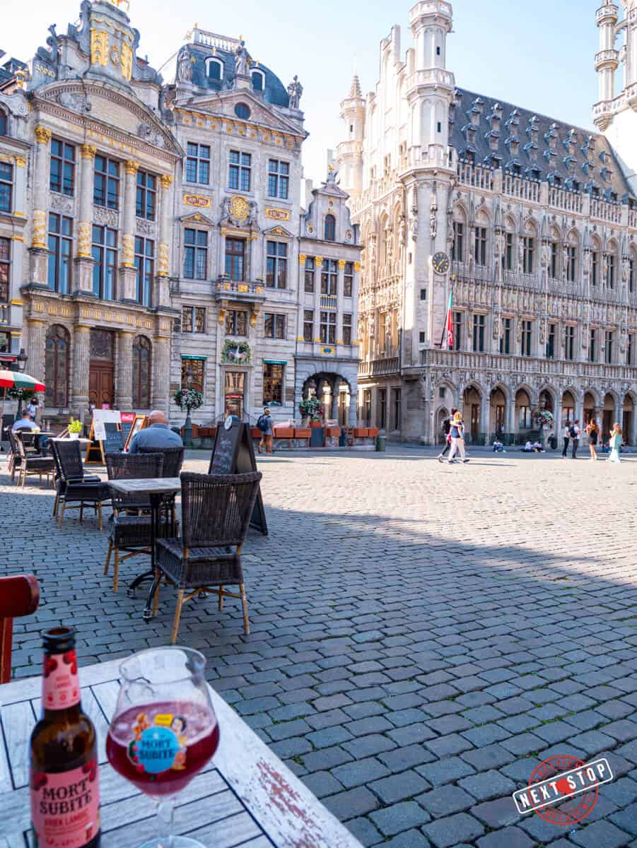 A view of Brussels' Grand Place with ornate historic buildings, a café table with a glass of Mort Subite beer in the foreground, and people walking in the distance.